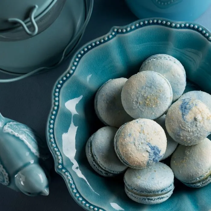 Top-down image of blue cotton candy french macaron cookies in a blue bowl next to a blue glass bird and blue candle lamp.